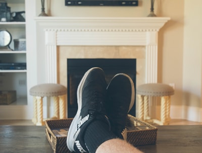 A person is relaxing in a living room with feet up on a wooden table. A TV mounted above a fireplace is displaying a popular streaming platform. Shelves on either side of the fireplace hold decorative items. A smartphone can be seen on the table.